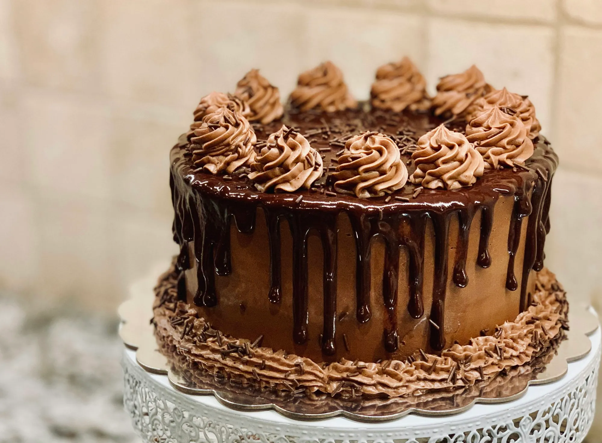 A chocolate drip cake topped with chocolate buttercream swirls on a white cake stand